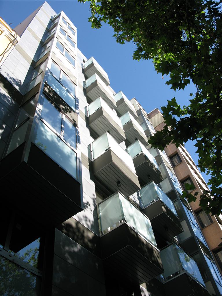 Edificio residencial de diseño contemporáneo con fachadas de vidrio y balcones flotantes. La estructura vertical maximiza el espacio en un entorno urbano, utilizando grandes ventanas y barandales de cristal que aportan modernidad y luminosidad a las viviendas. El uso de materiales como el acero y el vidrio realza el carácter minimalista del proyecto, creando un equilibrio entre funcionalidad y estética en construcciones urbanas de alta densidad